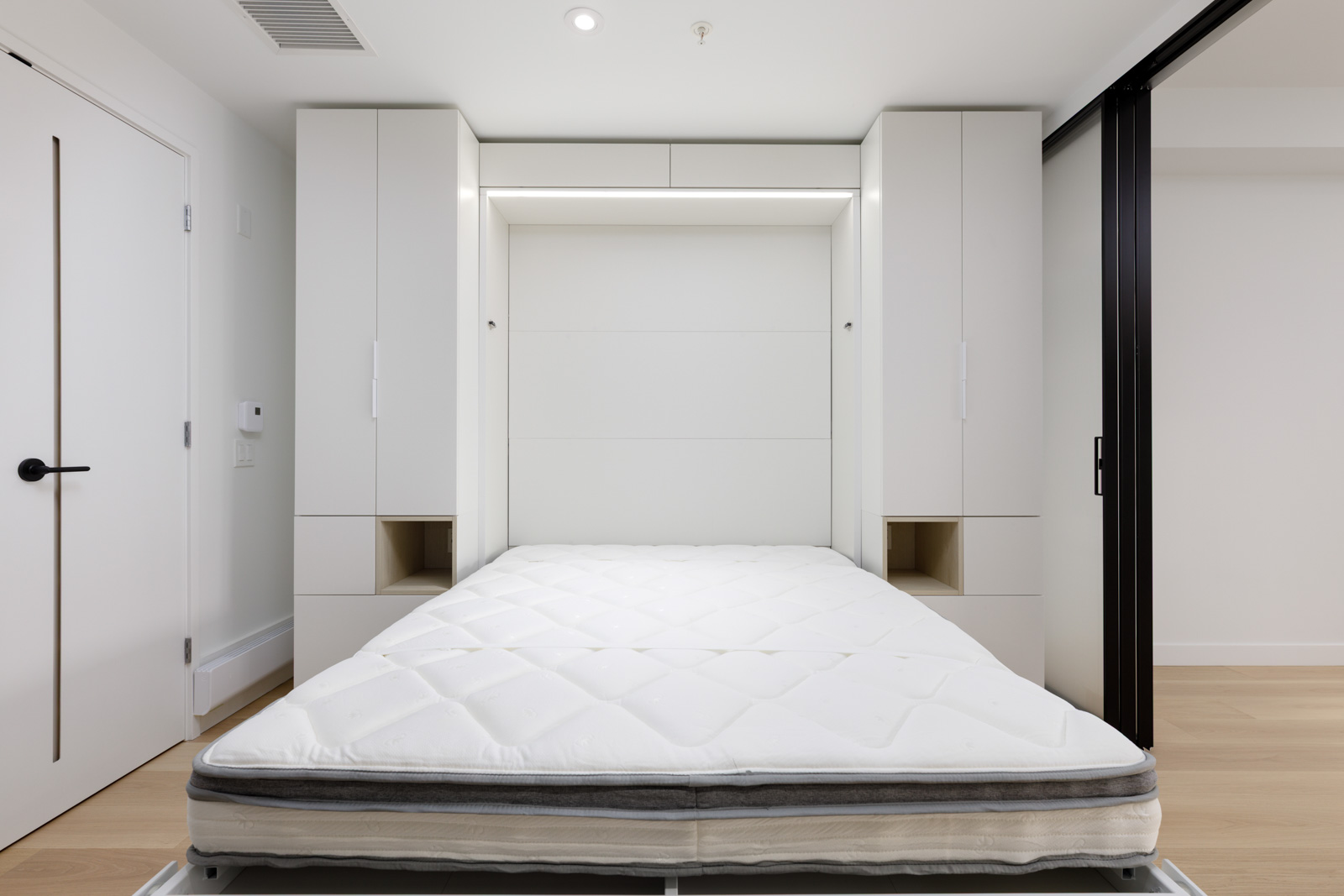 A room with a white Murphy bed pulled down, built-in cabinets on both sides, and light wood flooring.