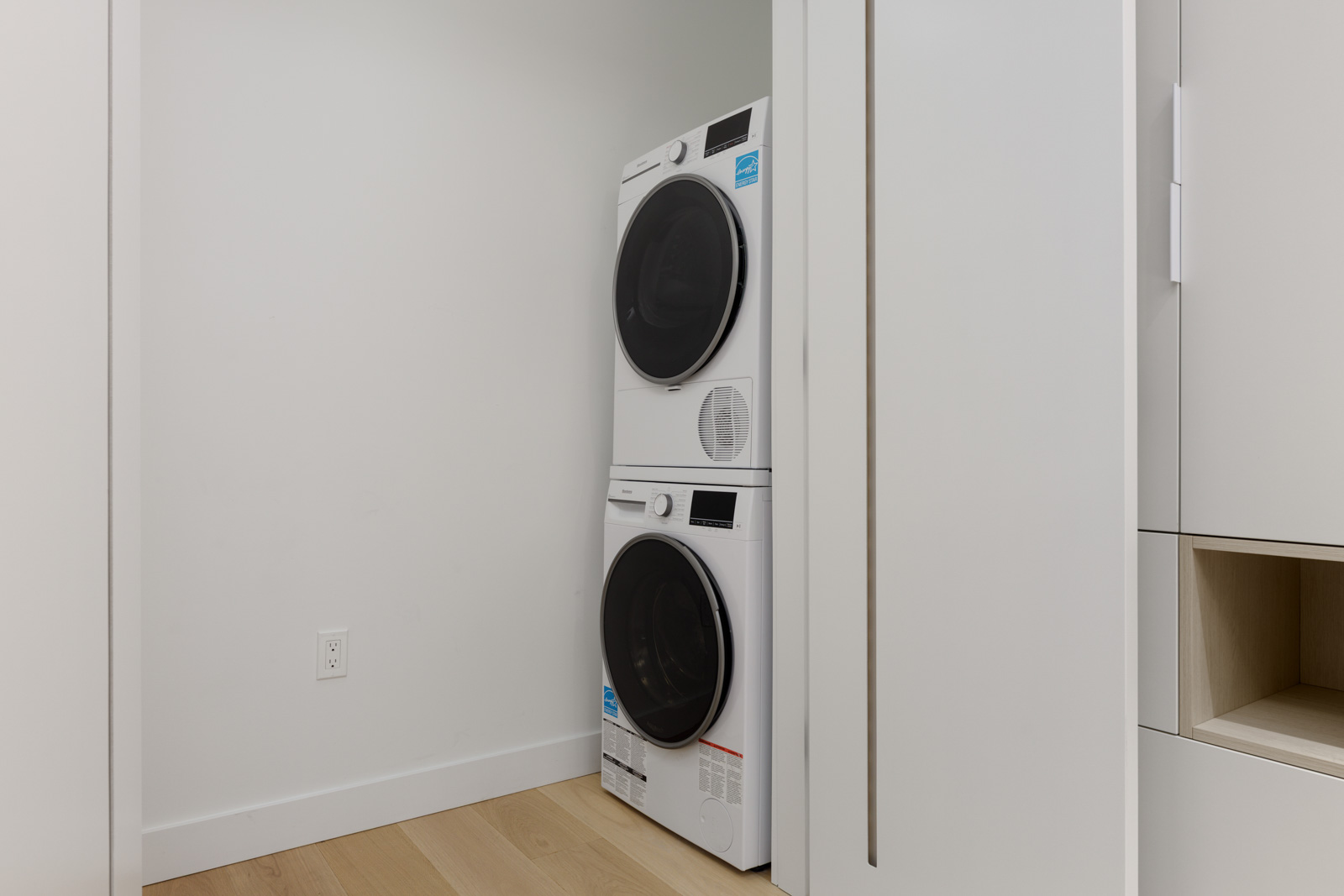 Stacked washer and dryer units in a small, modern laundry closet with light wood flooring and white walls.