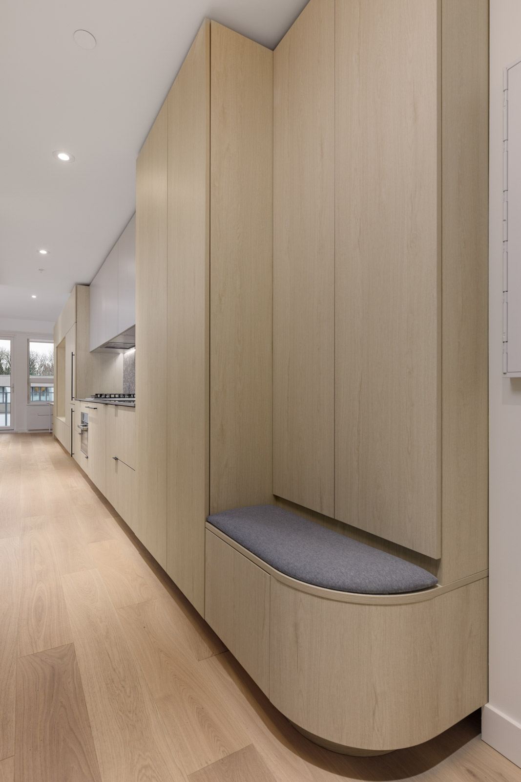 Modern kitchen with light wood cabinetry, integrated appliances, and a built-in cushioned bench. The space features light wood flooring and recessed ceiling lights.