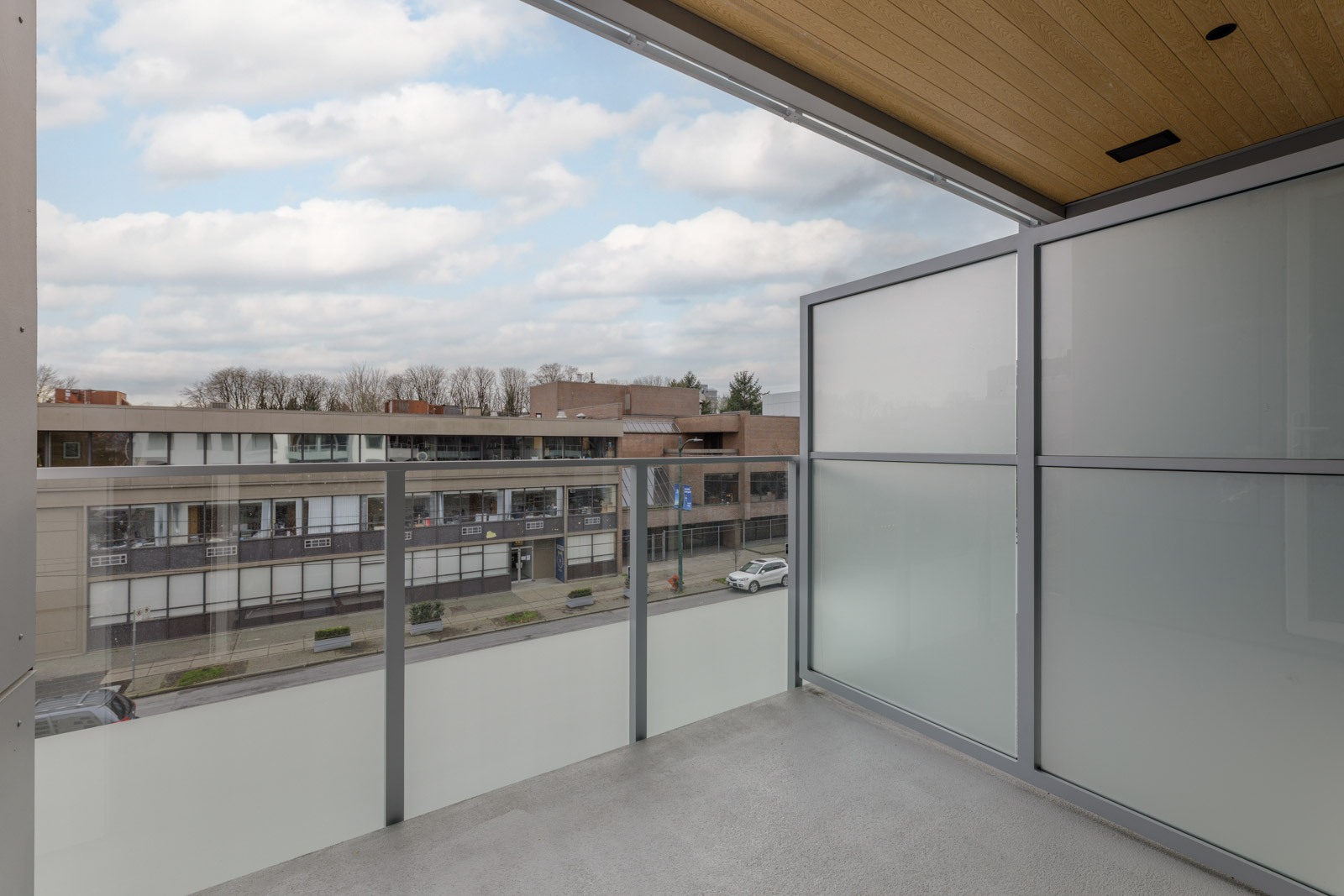 A modern balcony with frosted glass panels overlooks a street with low-rise buildings and parked cars under a partly cloudy sky.