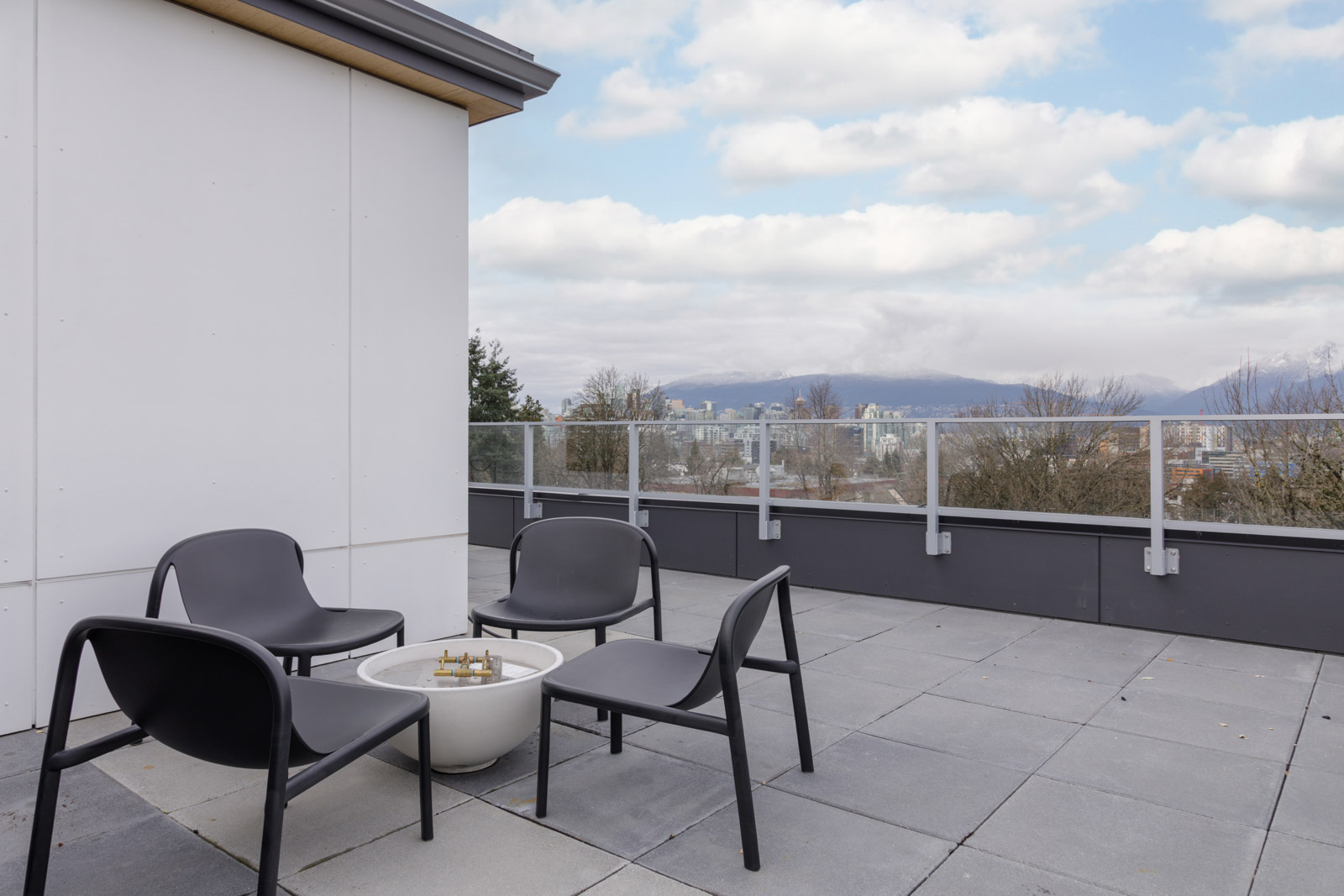 Rooftop patio with four black chairs around a fire pit, glass railing, and a cityscape with mountains in the background.