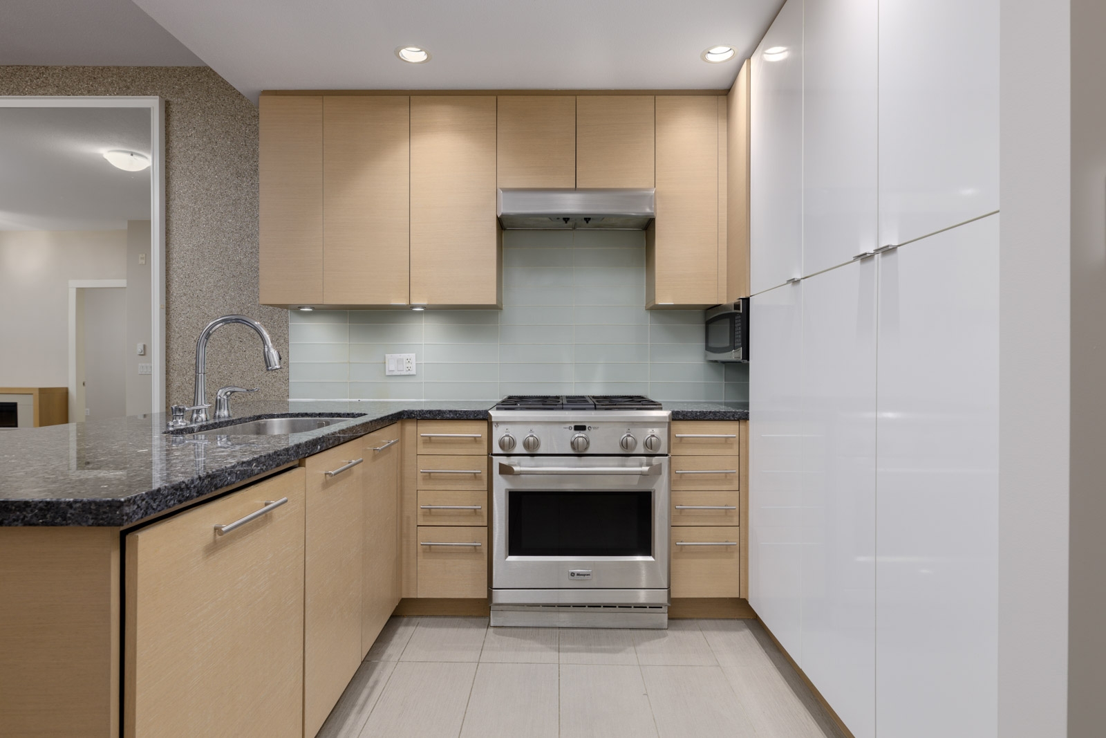 Modern kitchen with light wood cabinets, white pantry doors, stainless steel gas stove, black granite countertops, and a tile backsplash.