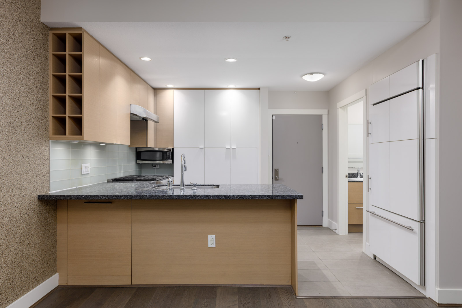 Modern kitchen with light wood cabinets, white upper cabinets, a black granite countertop, built-in appliances, and an entry door in the background.