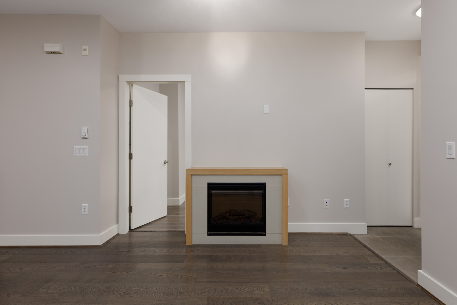 A minimalist room with light-colored walls, a dark wood floor, an electric fireplace, an open door, and a closed closet door.