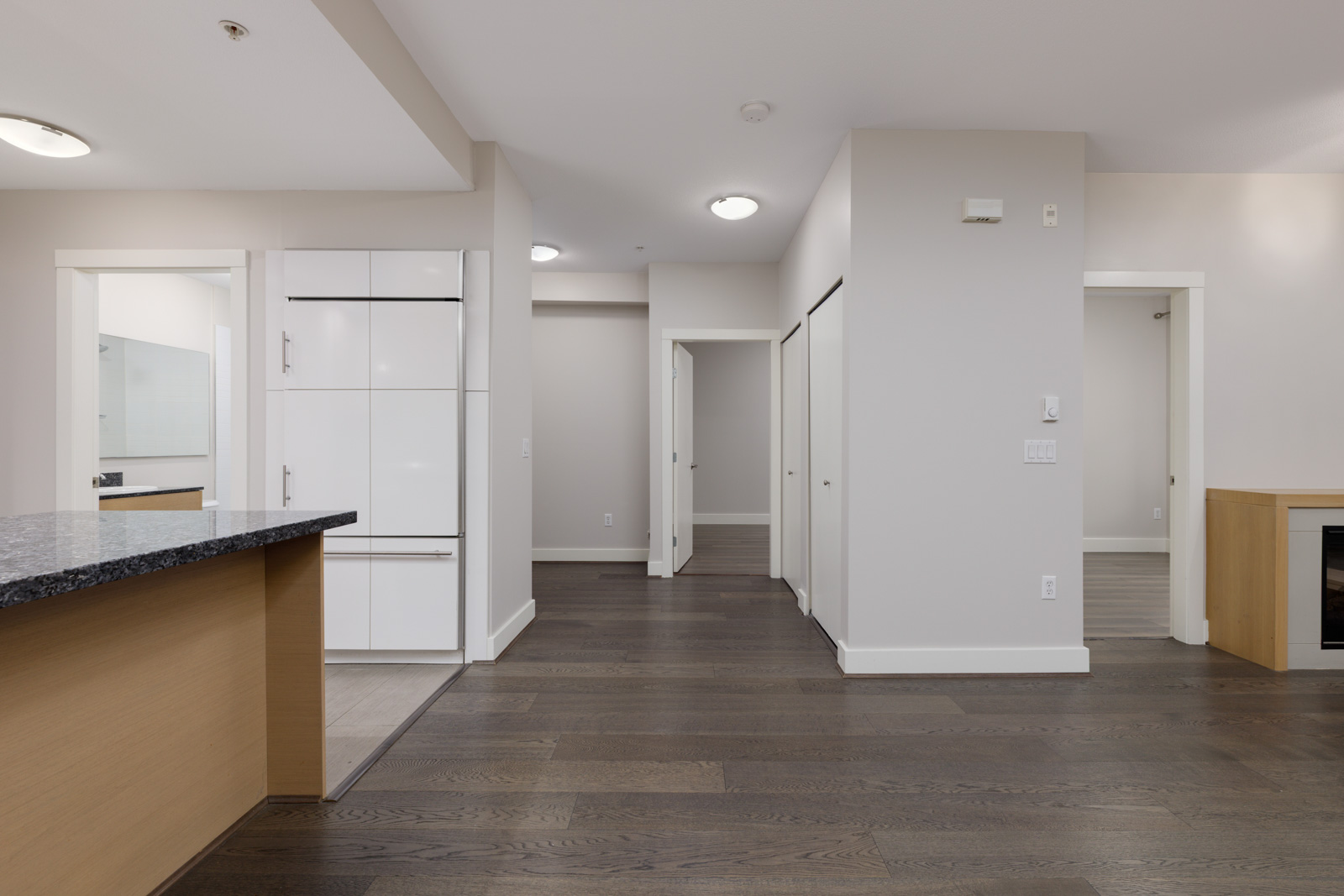 Interior view of a modern apartment with dark wood floors, white walls, multiple doorways, and a kitchen countertop on the left.