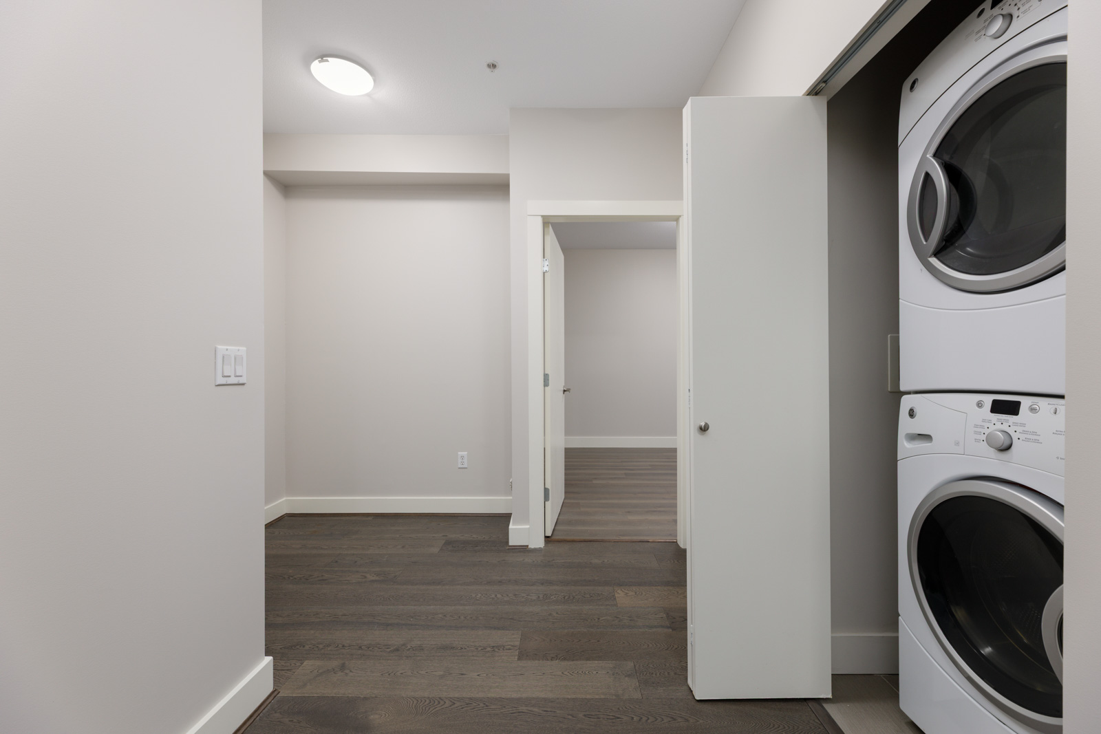 A modern laundry area with stacked washer and dryer behind a white closet door in a hallway with light walls and dark wood flooring.