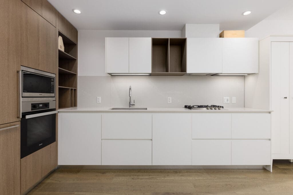 Modern kitchen with white cabinets, built-in appliances, wood accents, sink, gas cooktop, and open shelving, set against a light-colored backsplash and wood flooring.