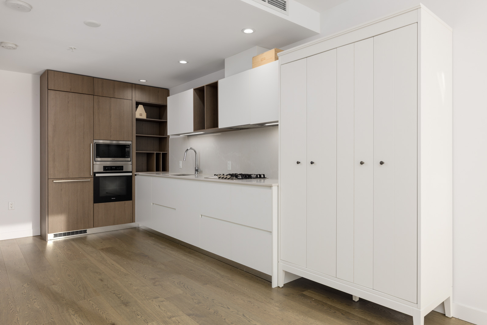Modern kitchen with wood flooring, built-in appliances, white cabinetry, open shelves, and a large white freestanding cabinet on the right.