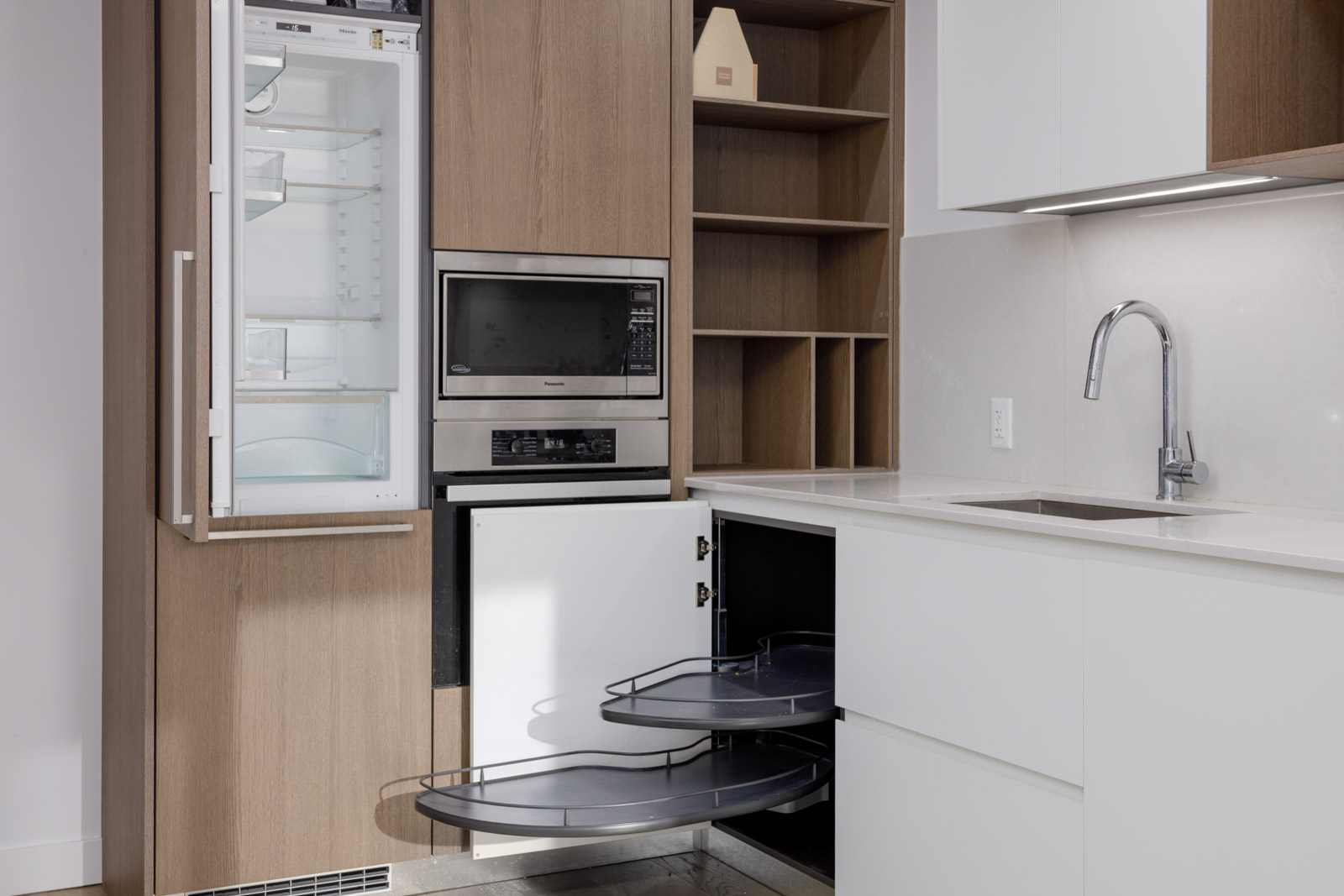 A modern kitchen with an open refrigerator, microwave, pull-out corner cabinet shelves, and a sink with a chrome faucet.