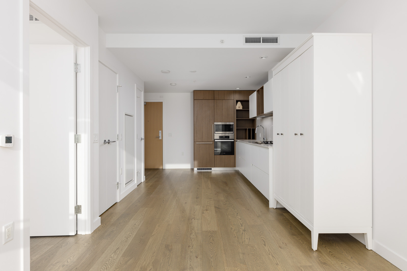 Modern kitchen with white and wood cabinetry, built-in oven, light wood flooring, and minimalistic design in a bright, unfurnished apartment.