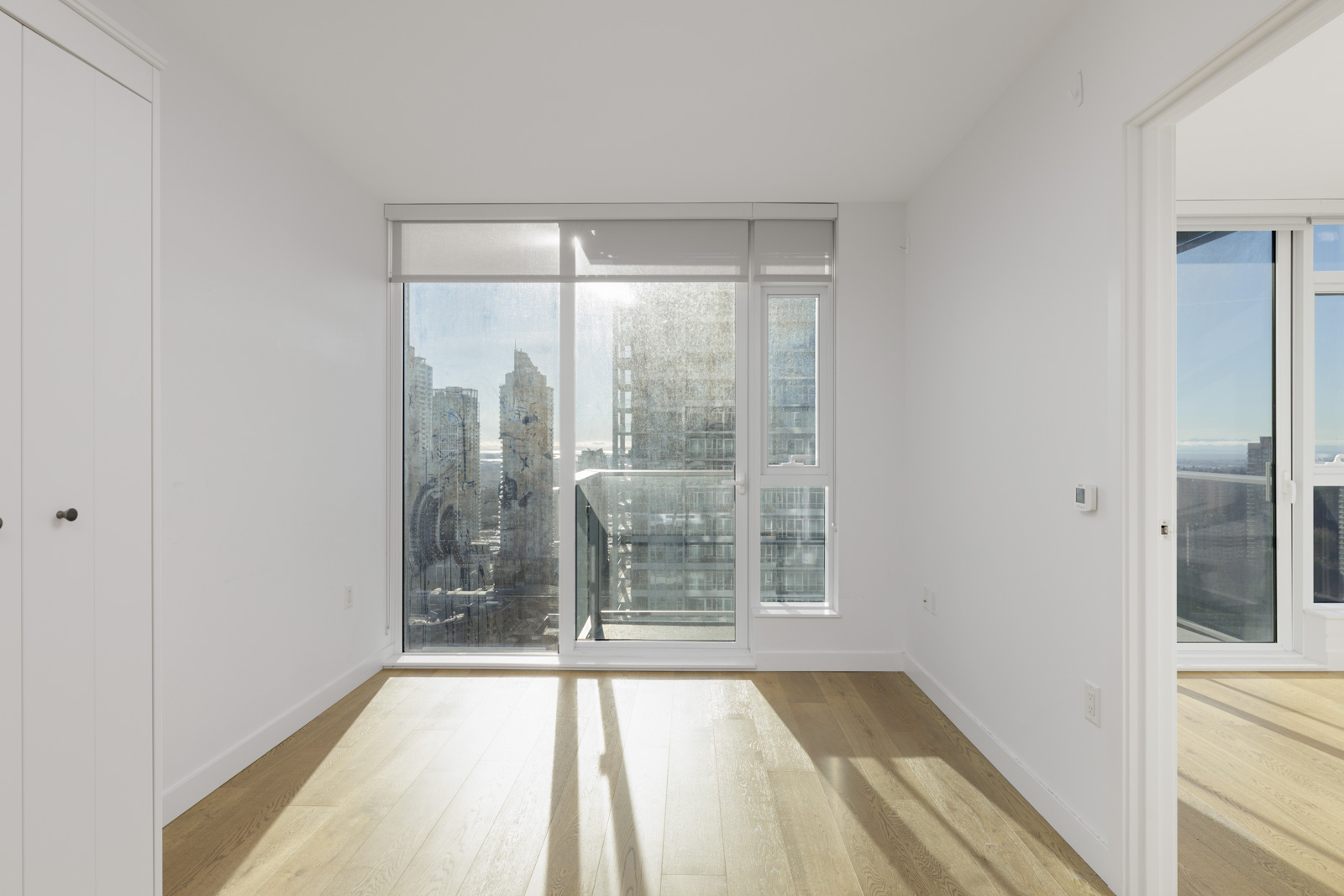Bright, empty room with light wood floors, large windows, and glass balcony door overlooking a city skyline on a sunny day.