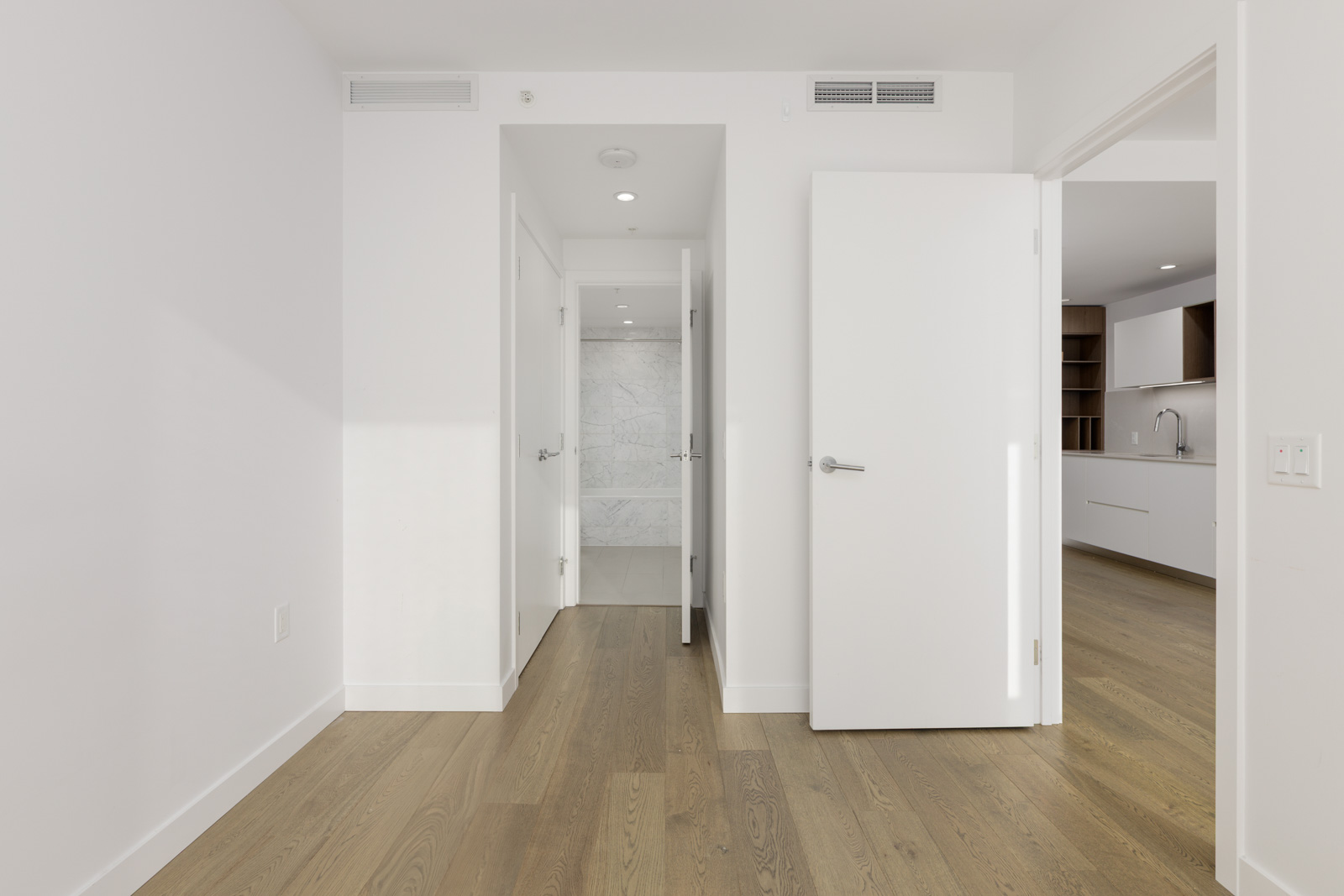 A minimal, modern interior with light wood flooring, white walls, three open doors, and a view into a kitchen and bathroom.