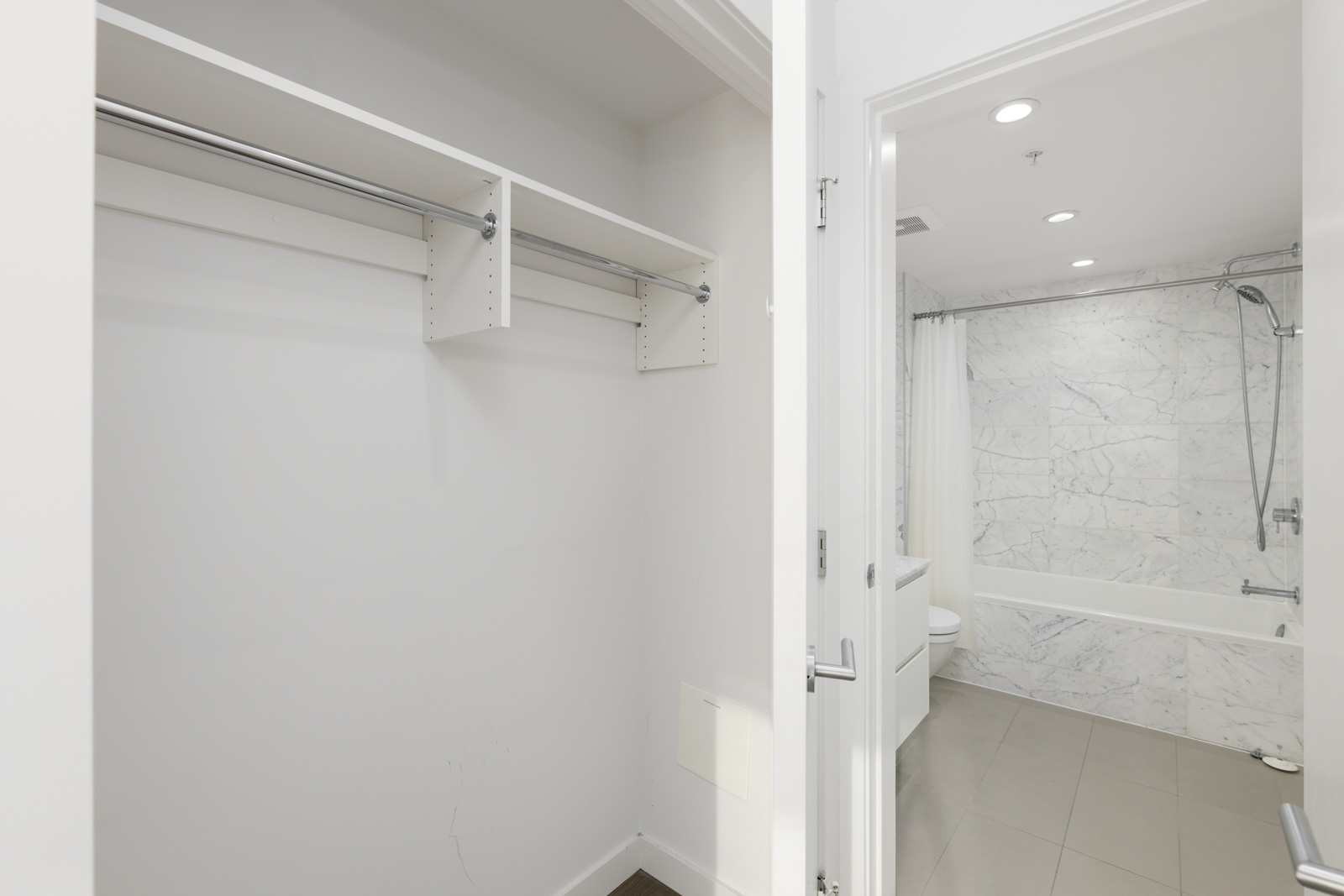 Empty white walk-in closet with metal hanging rods and shelves, next to a bathroom with a tiled shower and bathtub visible through an open door.