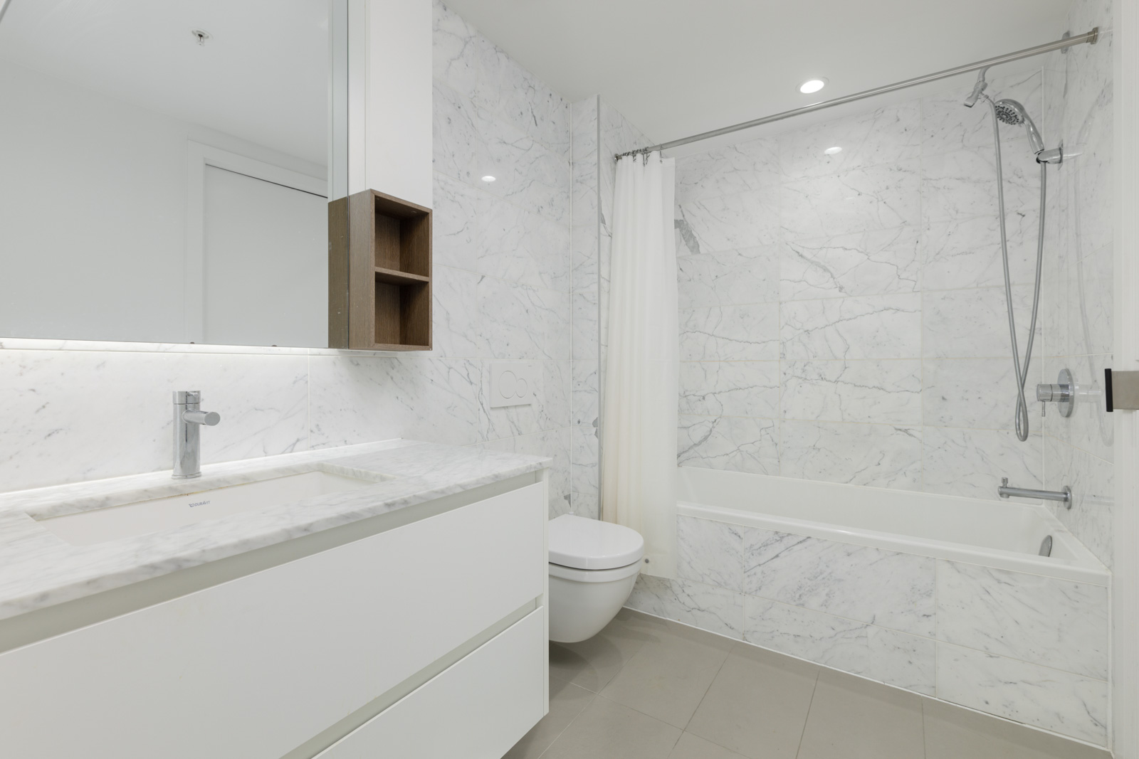 Modern bathroom with marble walls, a white vanity, wall-mounted toilet, bathtub with shower, and a white shower curtain.