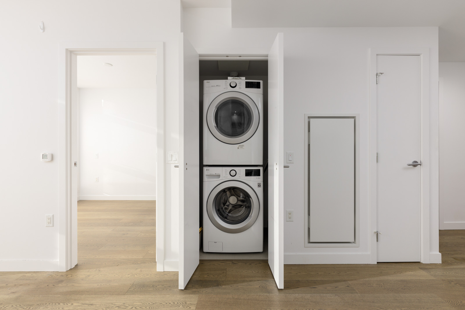 A stacked washer and dryer are installed in a small closet with open doors in a modern, minimalistic room with light wood floors and white walls.