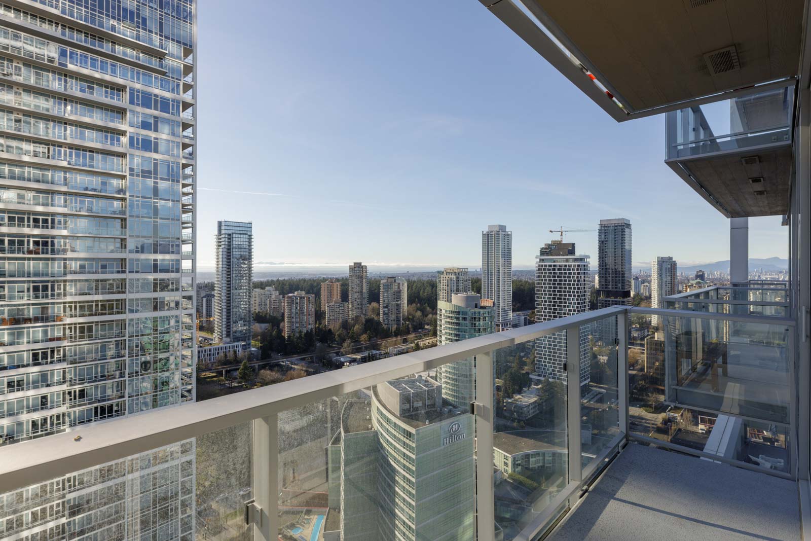 View from a high-rise balcony overlooking modern skyscrapers, trees, and distant water under a clear sky.