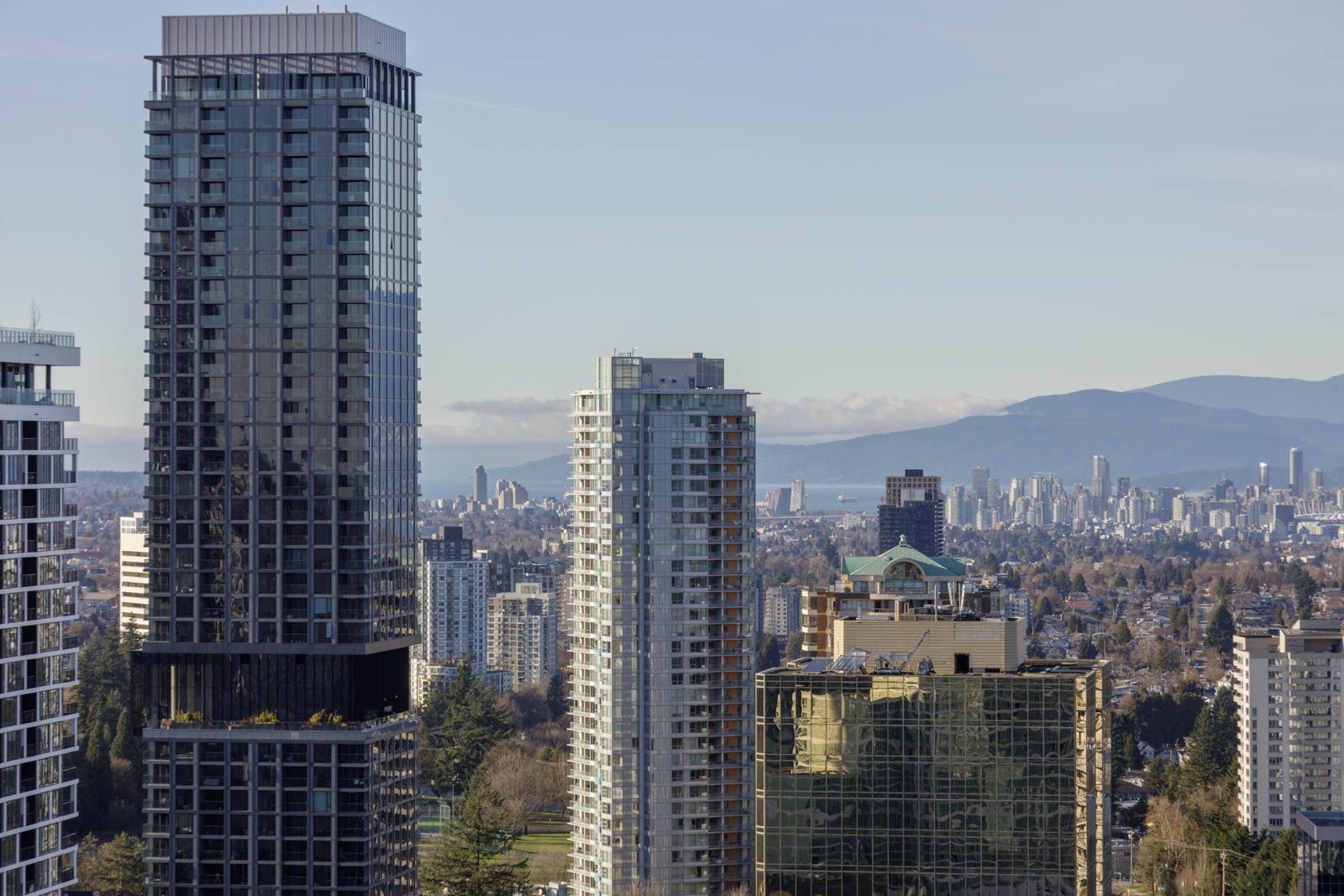 A cityscape with several tall modern buildings in the foreground and a distant view of mountains and more buildings under a clear sky.