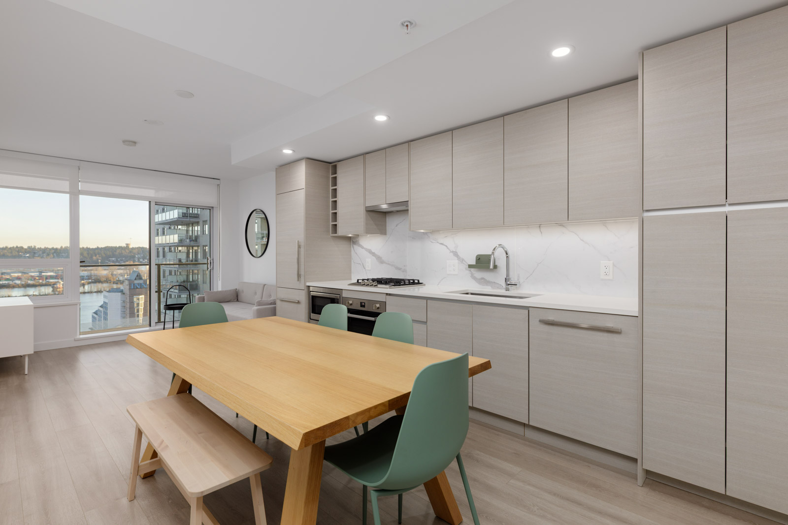 Modern kitchen and dining area with light wood cabinets, built-in appliances, a wooden table, green chairs, bench seating, and large windows with a city and water view.