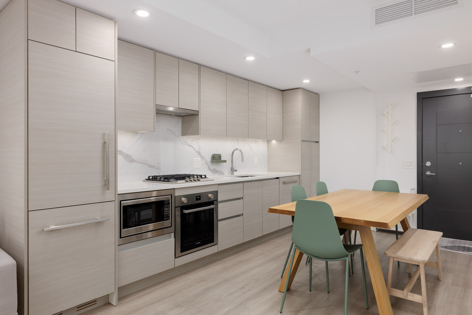 Modern kitchen with light wood cabinets, built-in appliances, a wooden dining table, green chairs, and a bench, featuring a marble backsplash and neutral decor.