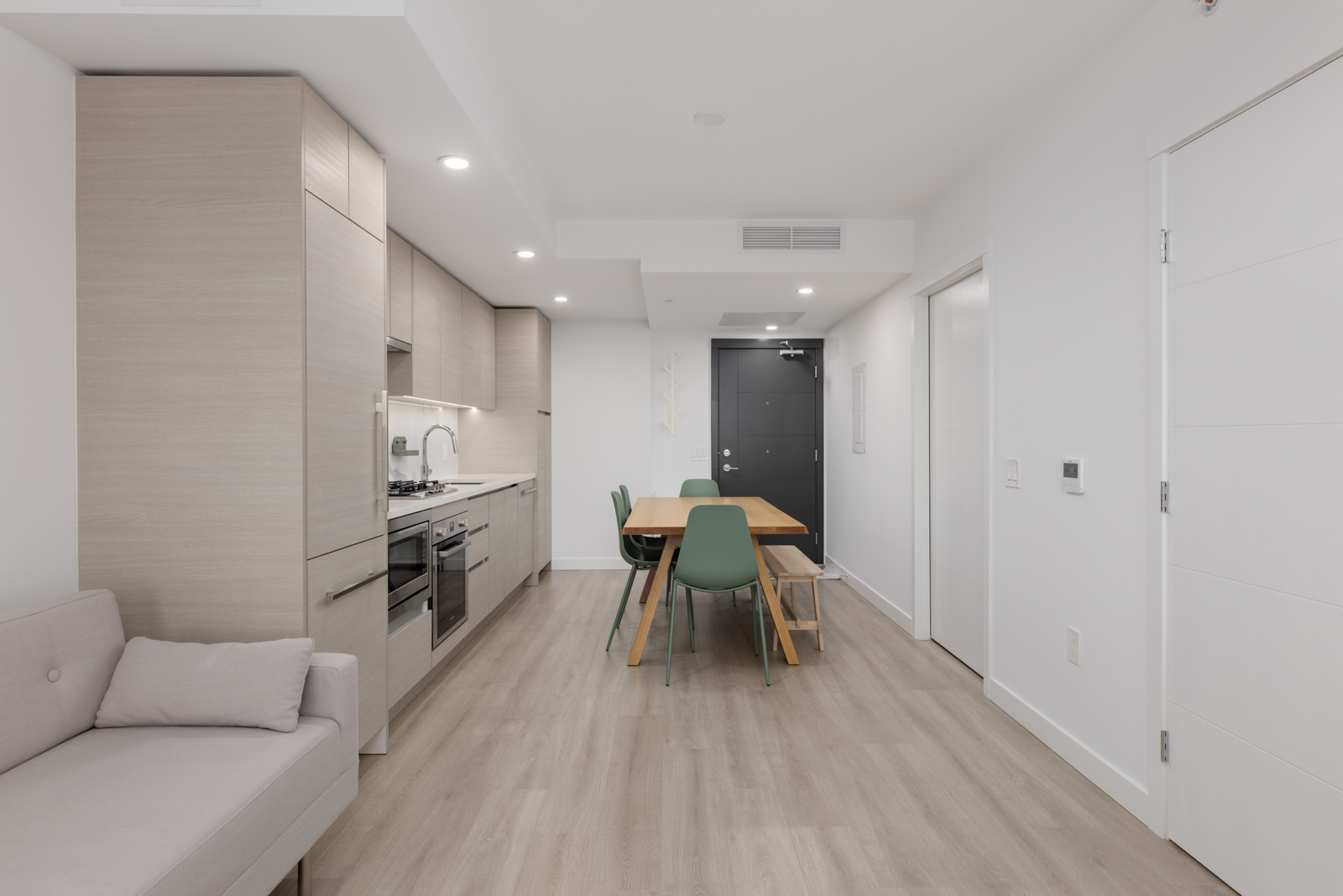 Modern apartment interior with light wood flooring, an open kitchen, dining table with four green chairs, and a gray sofa. White walls and ceiling with recessed lighting.