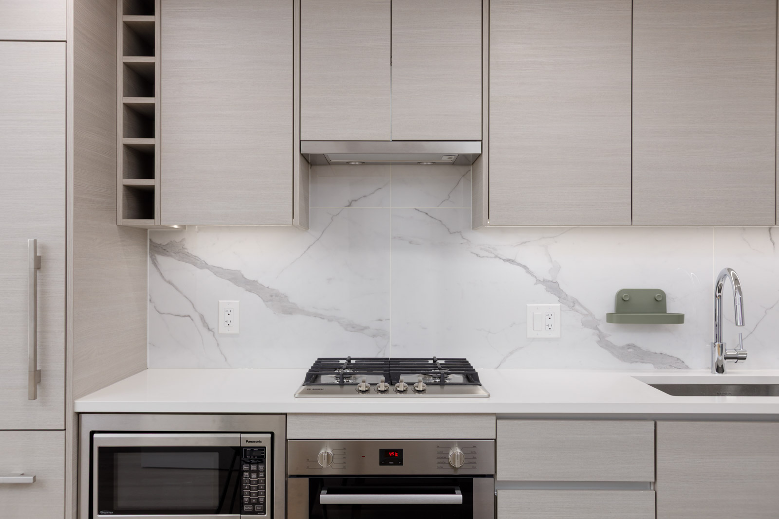 Modern kitchen with light-colored cabinets, built-in oven, gas stovetop, microwave, and marble backsplash, under-cabinet lighting, and a stainless steel sink with faucet.
