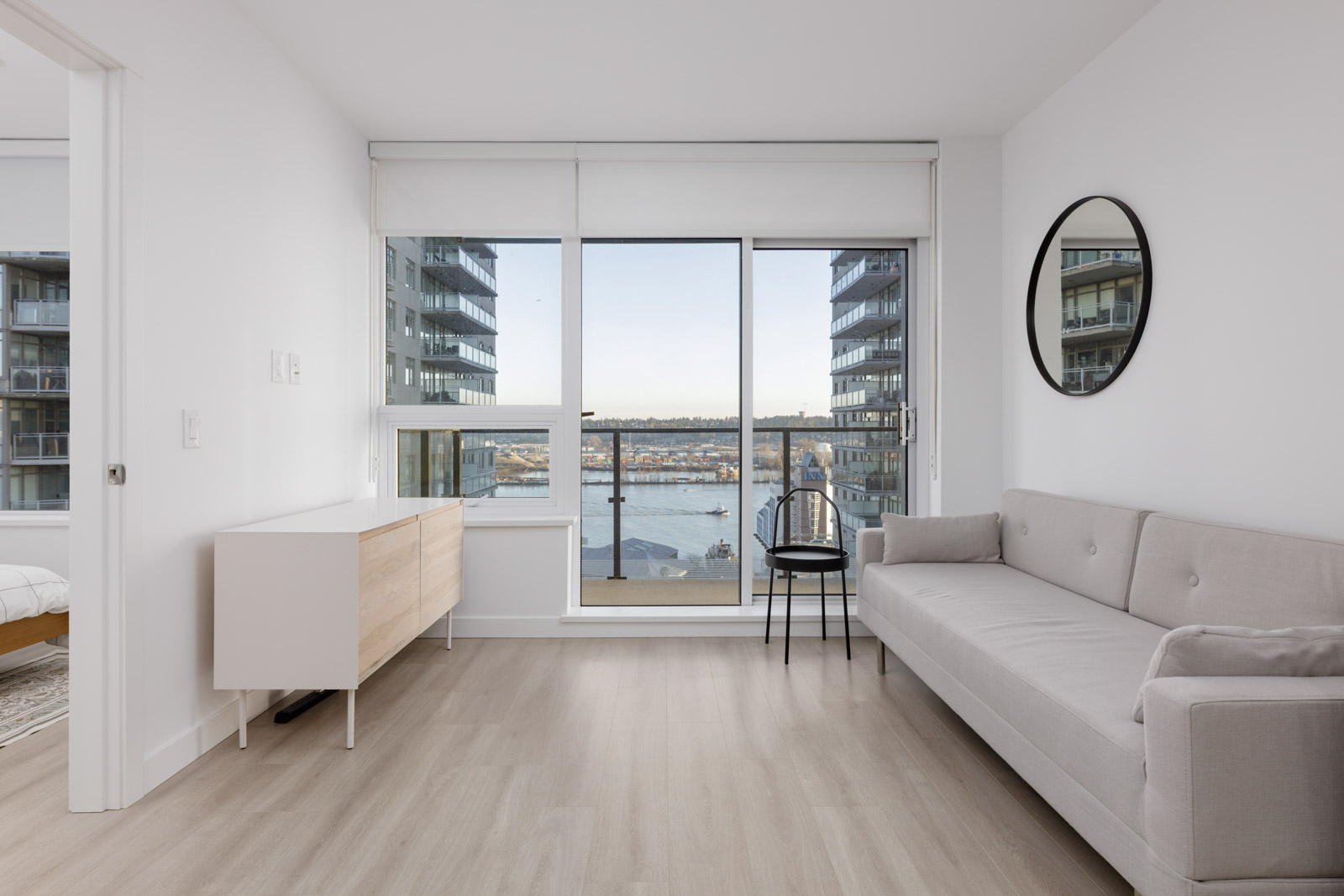 Modern minimalist living room with light wood floors, a gray sofa, a round mirror, and large windows overlooking a marina and apartment buildings.