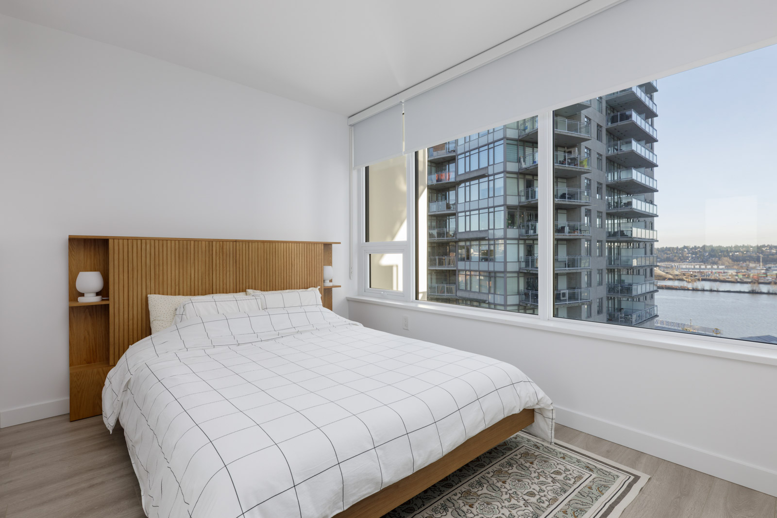 A modern bedroom with a wooden bed, white grid-patterned bedding, and a large window overlooking water and nearby apartment buildings.