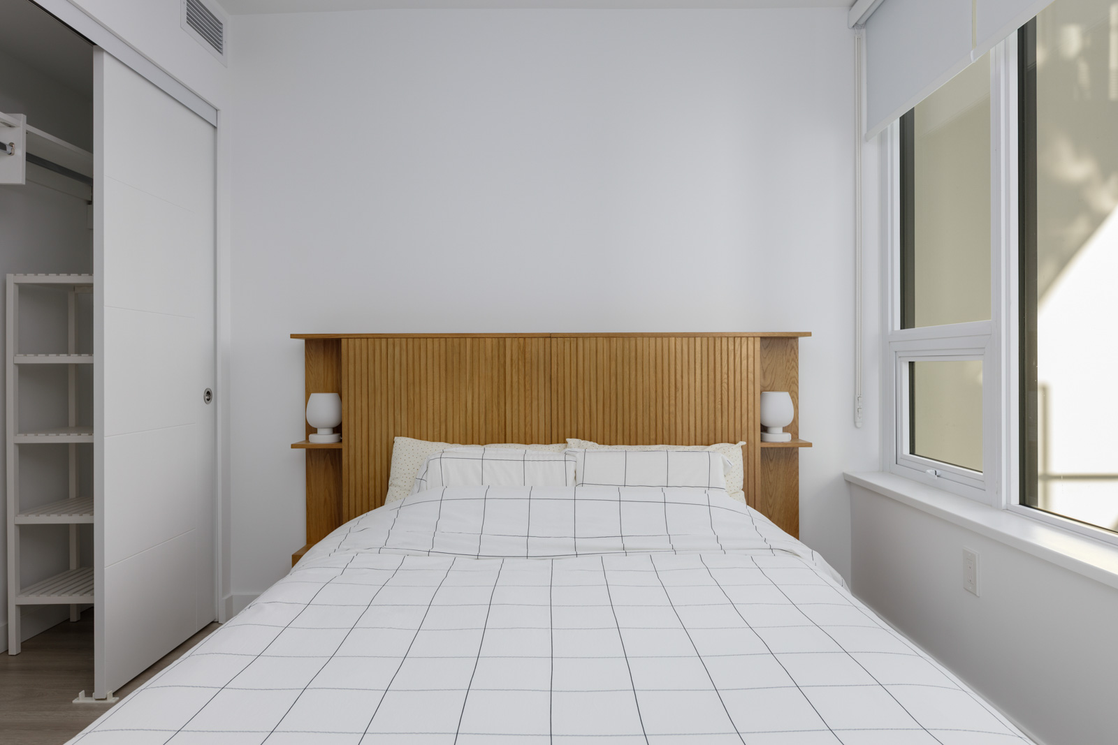 Minimalist bedroom with a wooden headboard, two pillows, a bed with a white grid-patterned duvet, two small lamps, and a window letting in natural light.