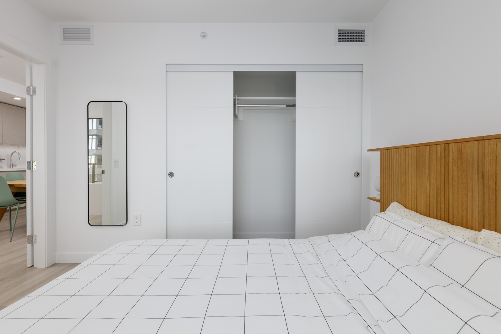 A modern bedroom with a grid-patterned bedspread, wooden headboard, built-in closet with sliding doors, wall mirror, and a view into a kitchen area.