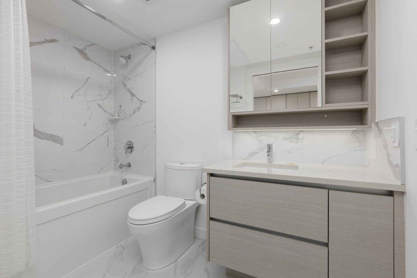 Modern bathroom with a bathtub-shower combo, toilet, light wood vanity, marble-patterned tiles, wall-mounted mirror cabinet, and shelves.