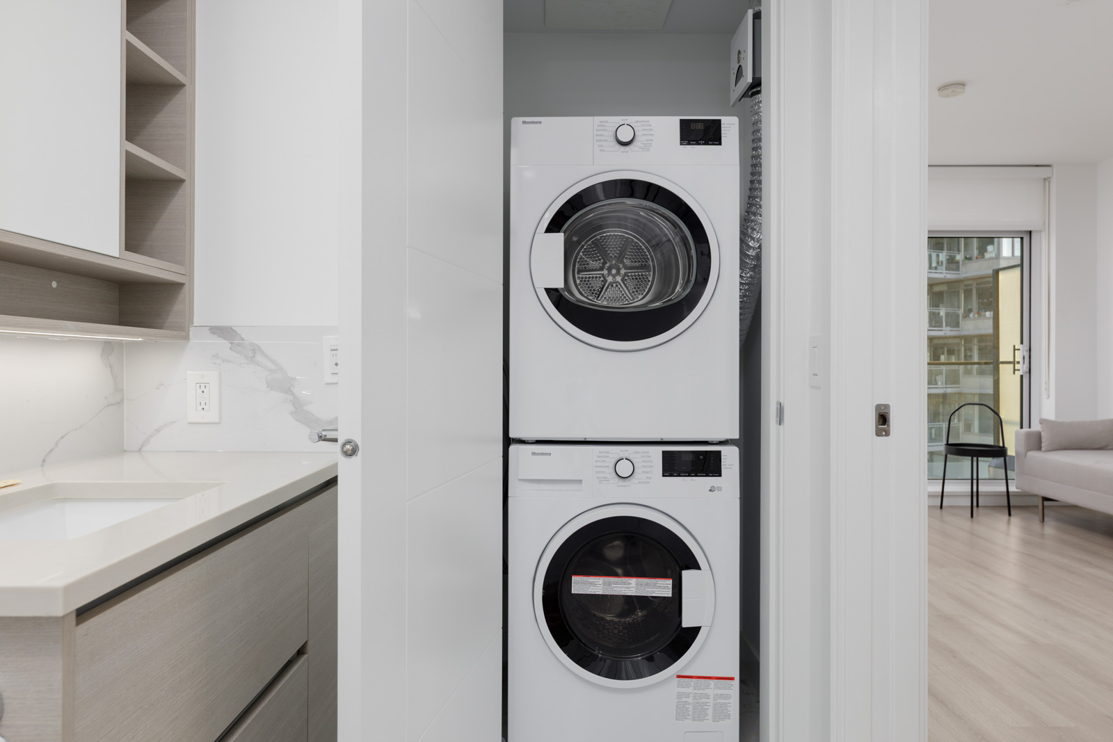 A stacked washer and dryer are installed in a narrow laundry closet next to a modern kitchen with light cabinetry and marble backsplash.