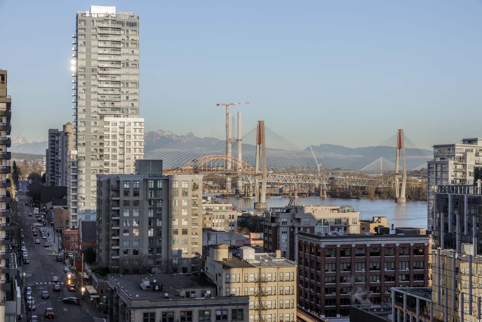 View of a city with mid-rise and high-rise buildings, a large cable-stayed bridge over a river, and mountains in the background under a clear sky.