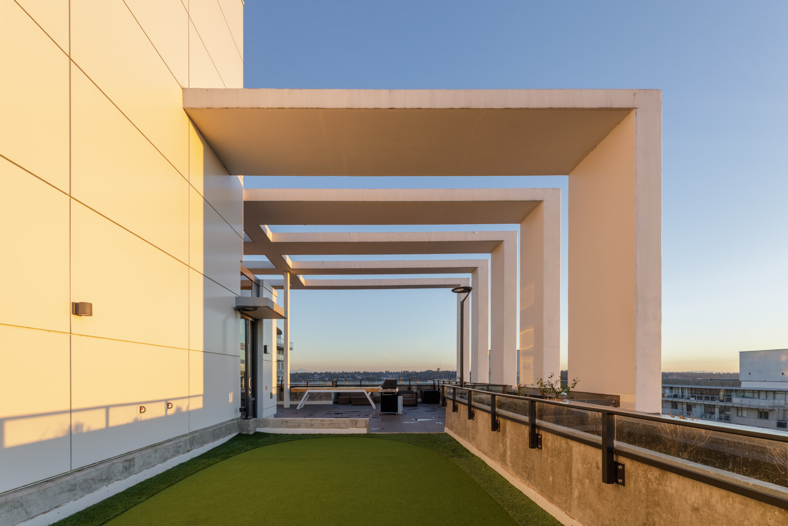 Modern rooftop with geometric white frames, green artificial turf, glass doors, and cityscape view under a clear sky at sunset.