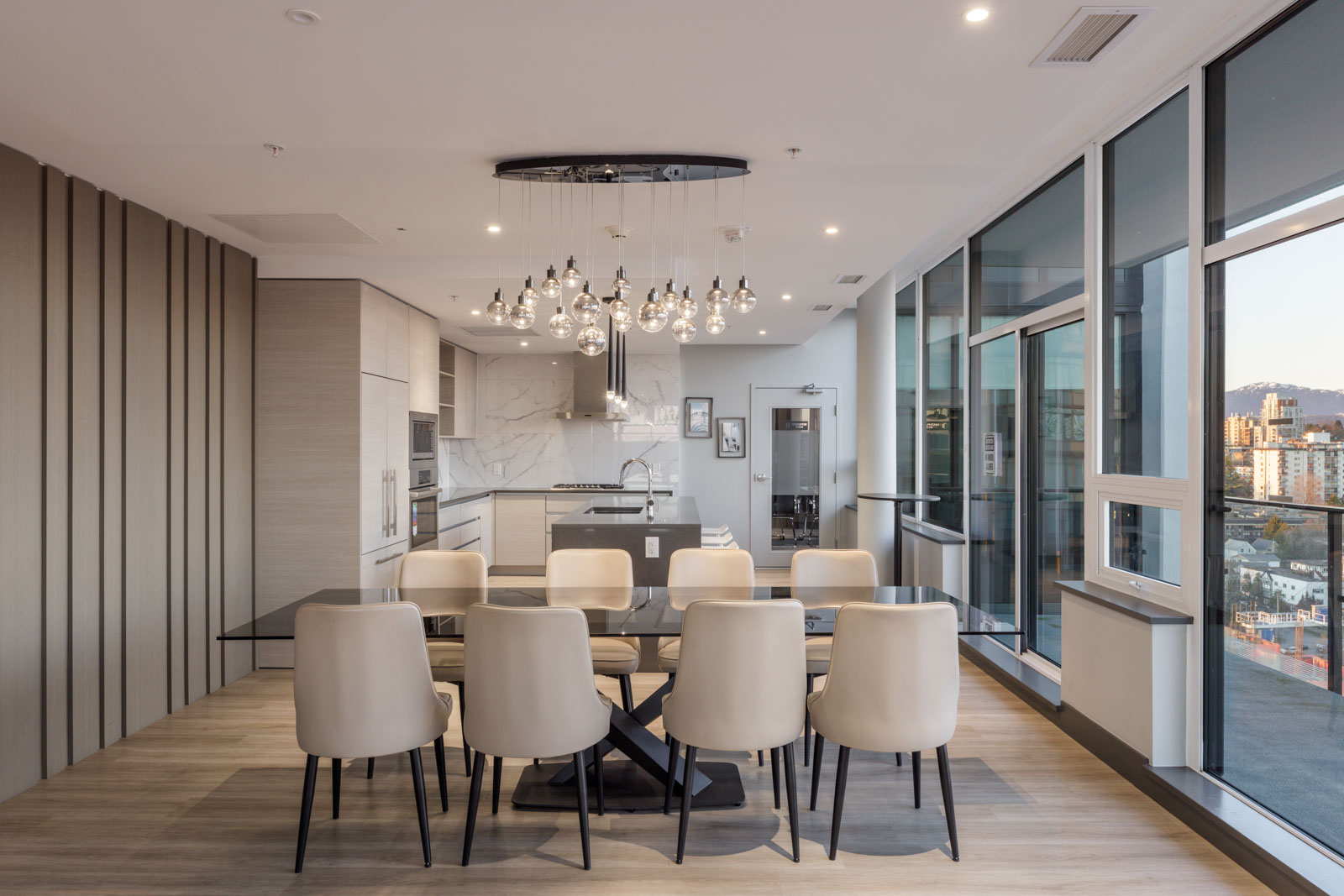 Modern dining area with a glass table and white chairs, open to a sleek kitchen and large windows offering a city view.