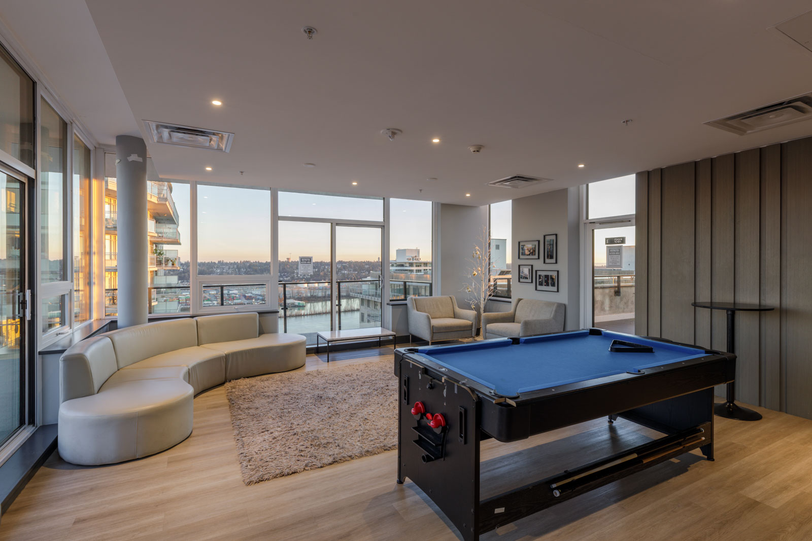 Modern lounge area with large windows, a curved white sofa, two armchairs, a rug, framed photos on the wall, and a pool table with a city view in the background.
