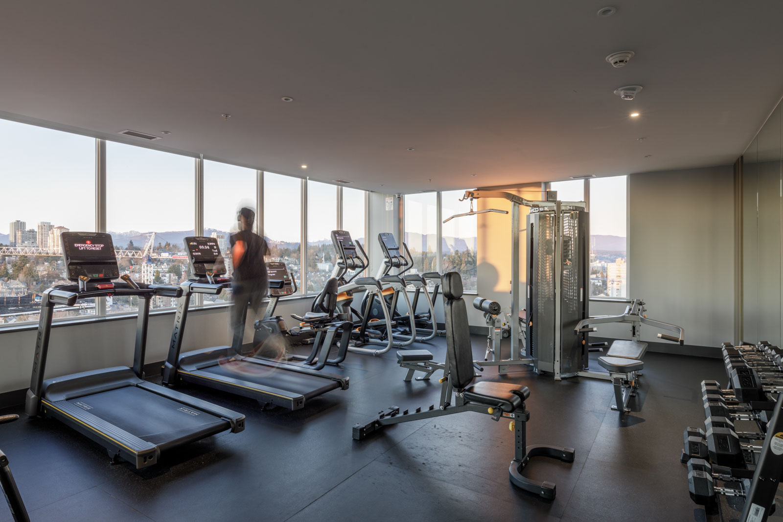 Modern gym with treadmills, ellipticals, weight machines, and dumbbells. Large windows provide a city view. One person is running on a treadmill.