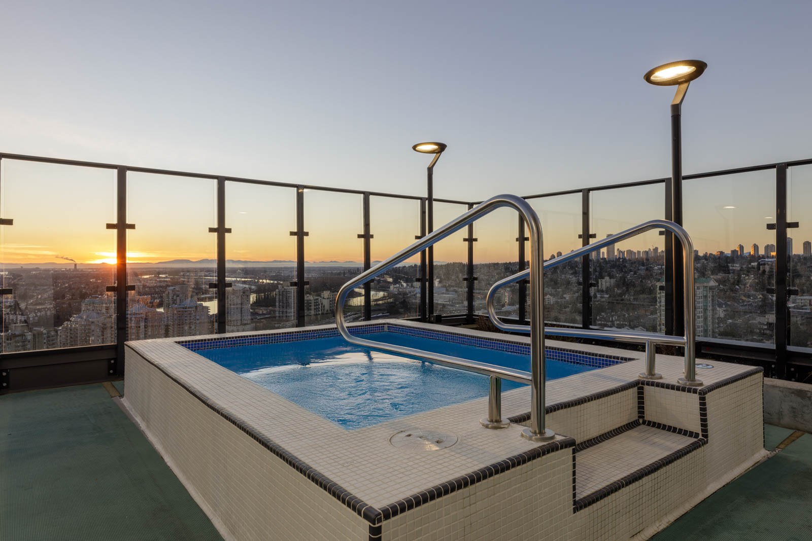 A rooftop hot tub with tiled steps and metal railings overlooks a city skyline at sunset, surrounded by glass barriers and outdoor lights.