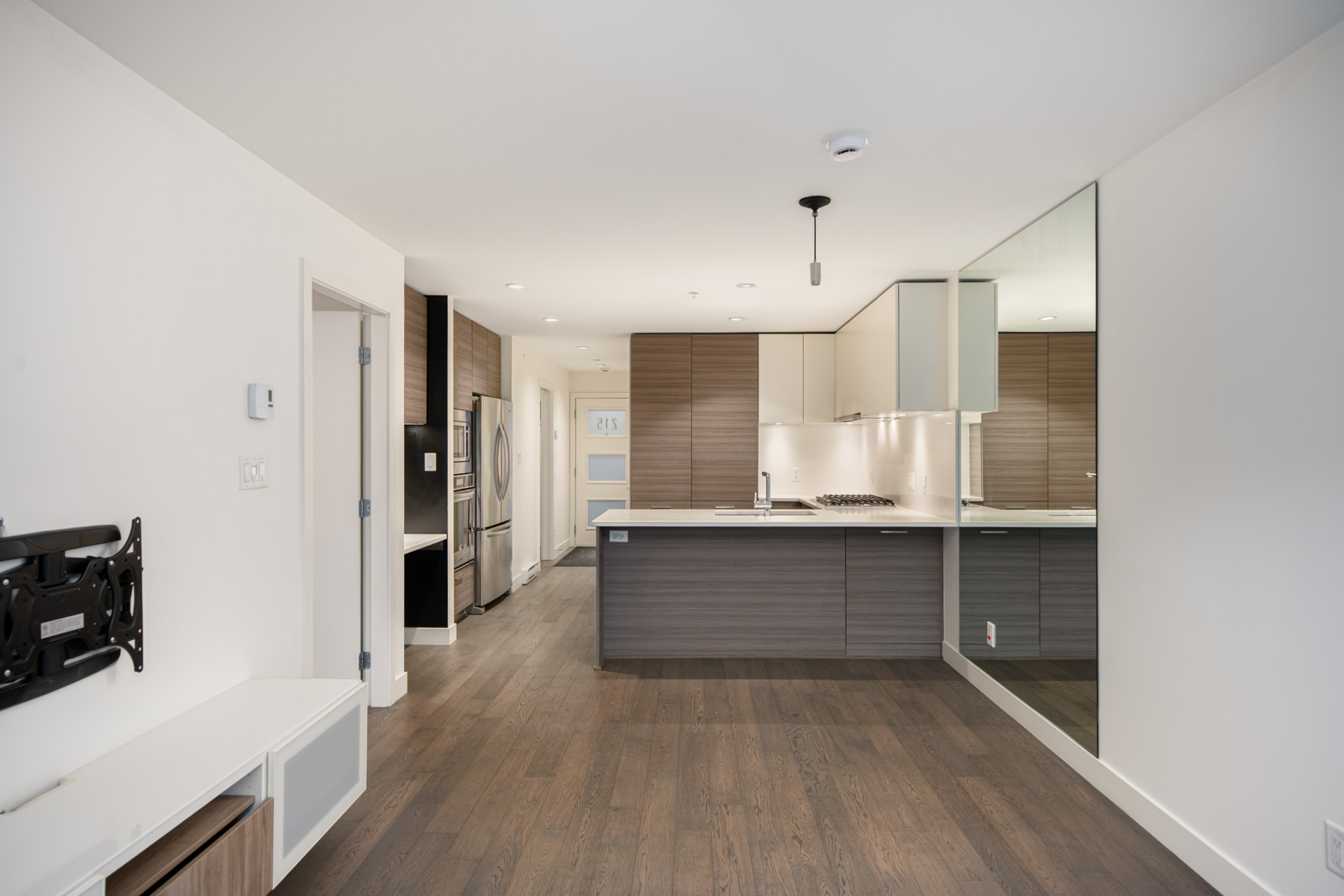 Modern kitchen with wooden floors, stainless steel appliances, brown cabinets, a large mirror on one wall, and a mounted TV bracket in the adjacent living area.