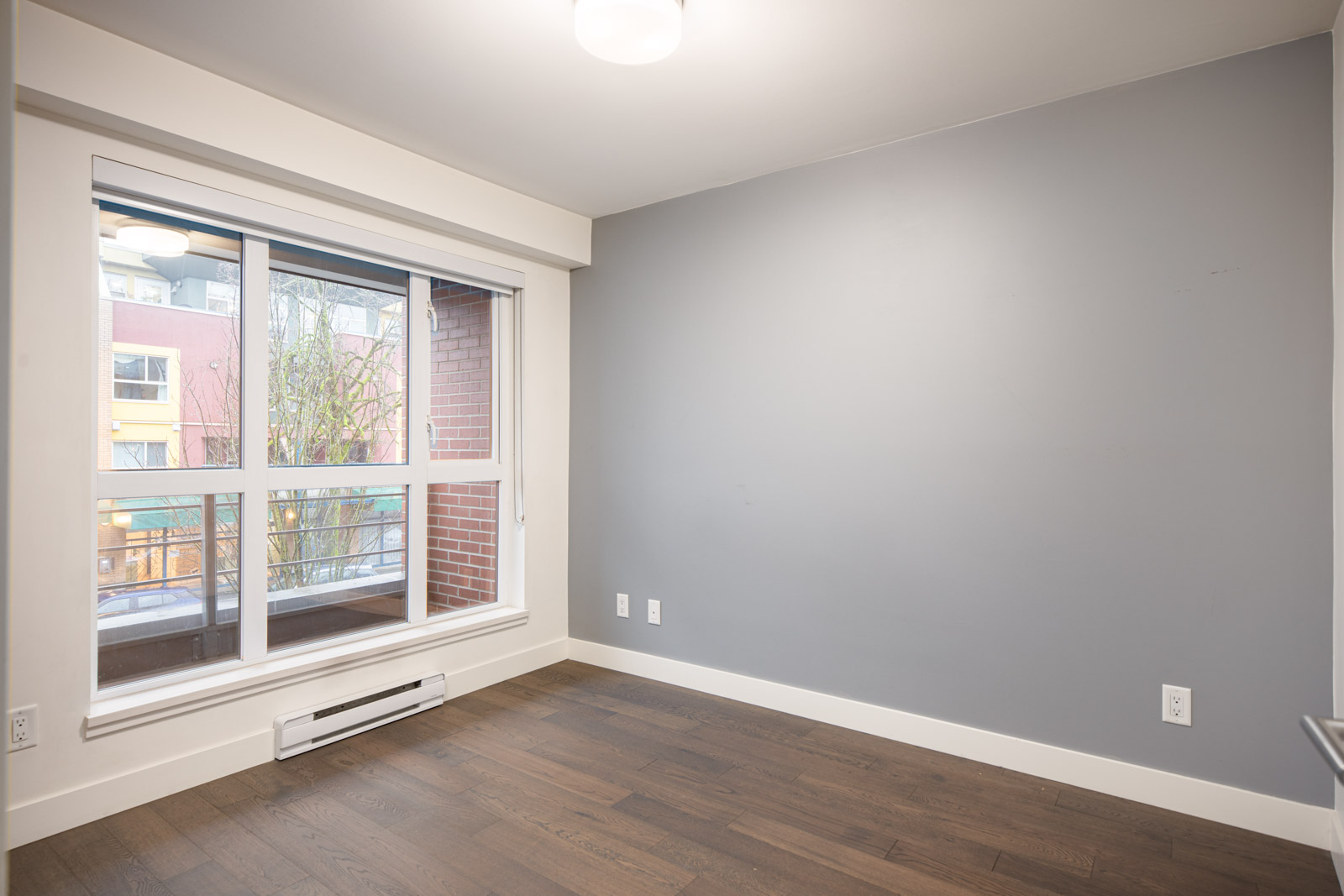 Empty room with gray walls, large window, white trim, baseboard heater, and dark wood flooring. Daylight is visible through the window.