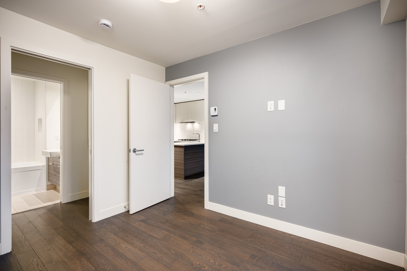 Empty room with dark wood floor, a gray accent wall, and an open door leading to a bathroom and kitchen area. Multiple power outlets and a thermostat are visible on the wall.