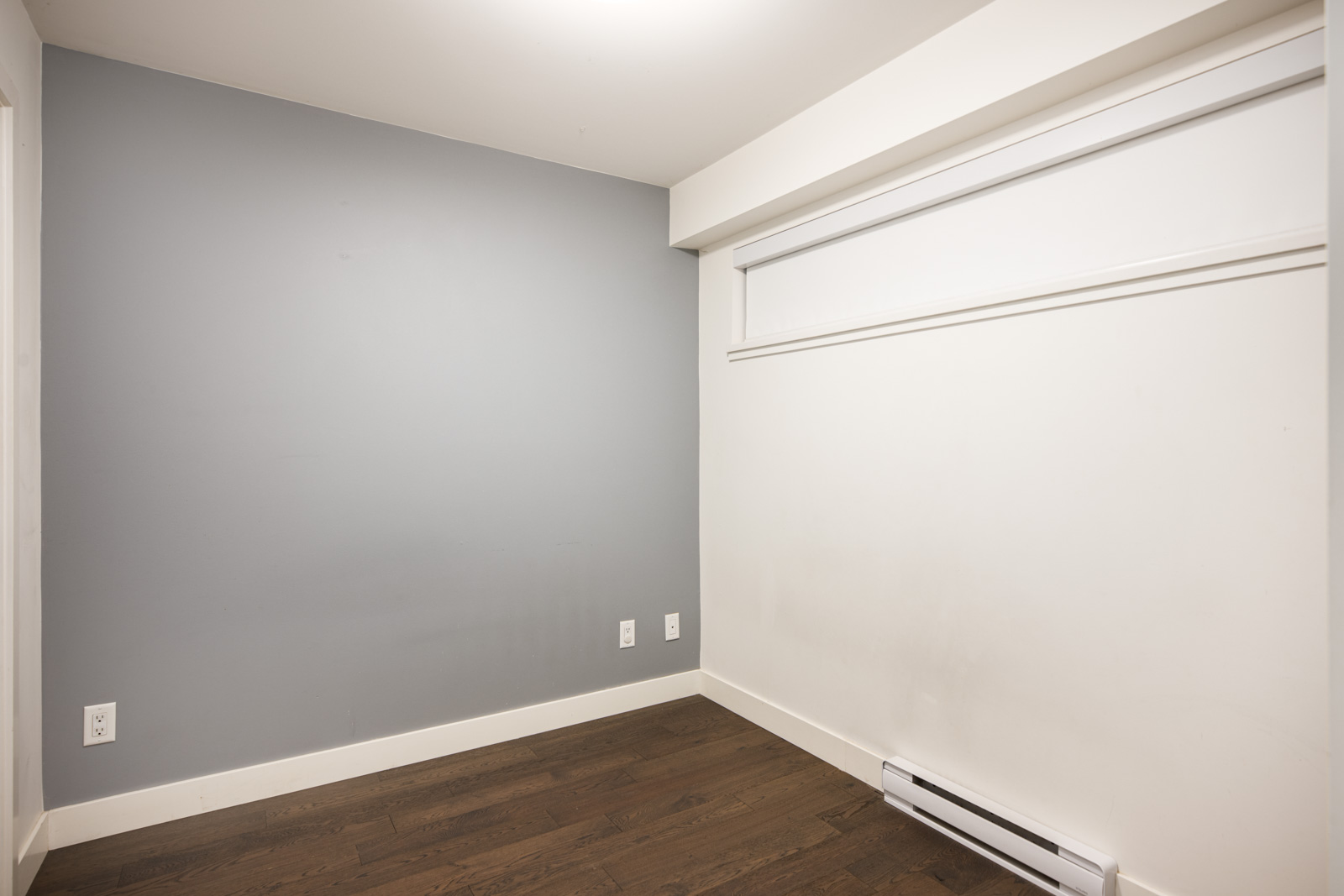 Empty room with light gray and white walls, dark wood flooring, a baseboard heater, electrical outlets, and a rectangular high window.