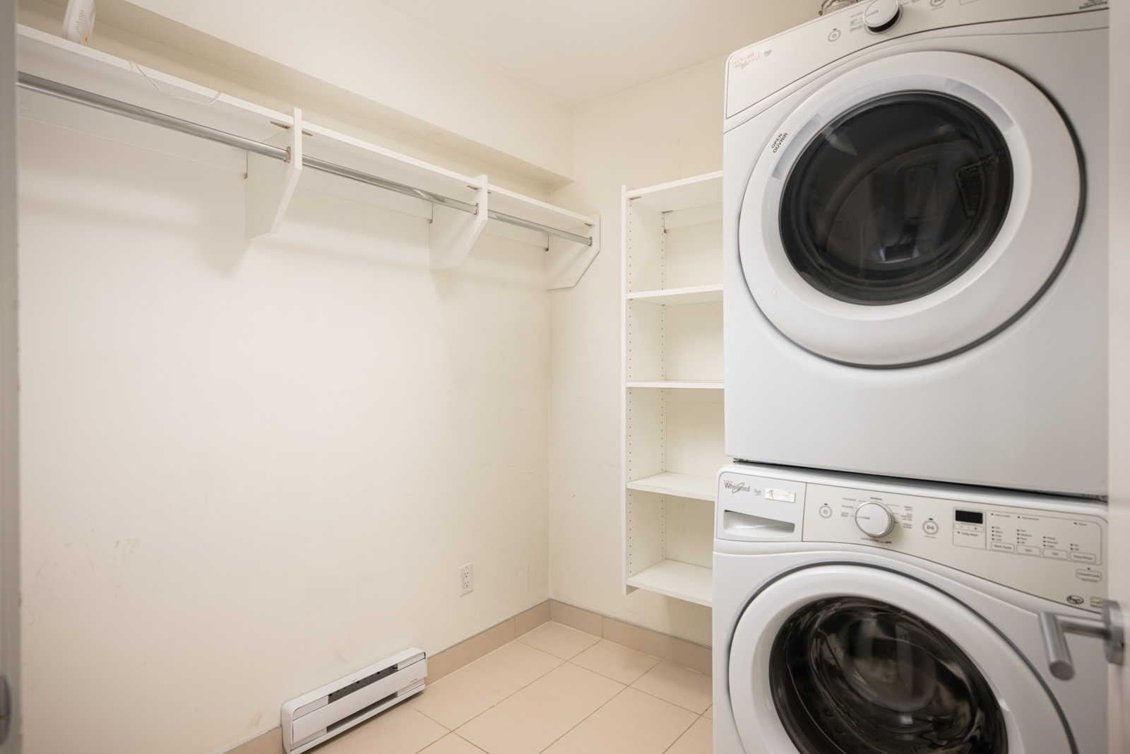 A laundry room with a stacked washer and dryer, wall shelving, open hanging rod, and tile flooring.