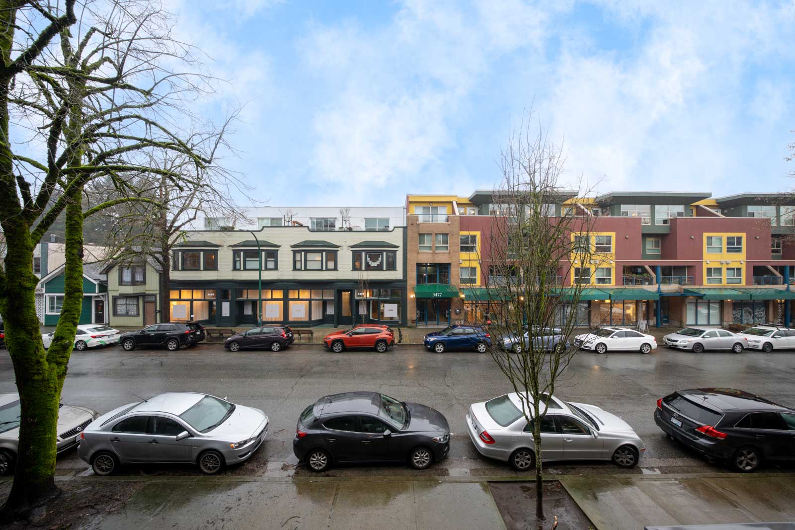 Street view of parked cars in front of colorful, modern low-rise buildings on a wet, overcast day. Leafless trees line the sidewalk.