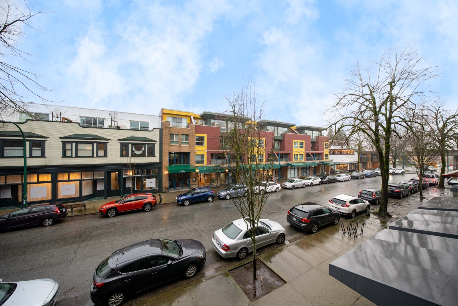 A street view of parked cars along a wet road in front of modern commercial and residential buildings on a cloudy day.