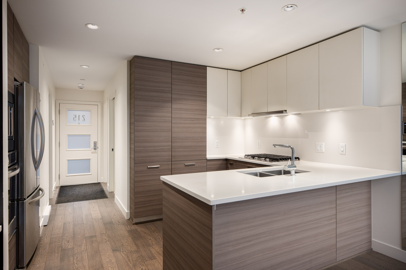 Modern kitchen with wood and white cabinetry, stainless steel appliances, white countertops, and a double sink, viewed from the entrance hallway.