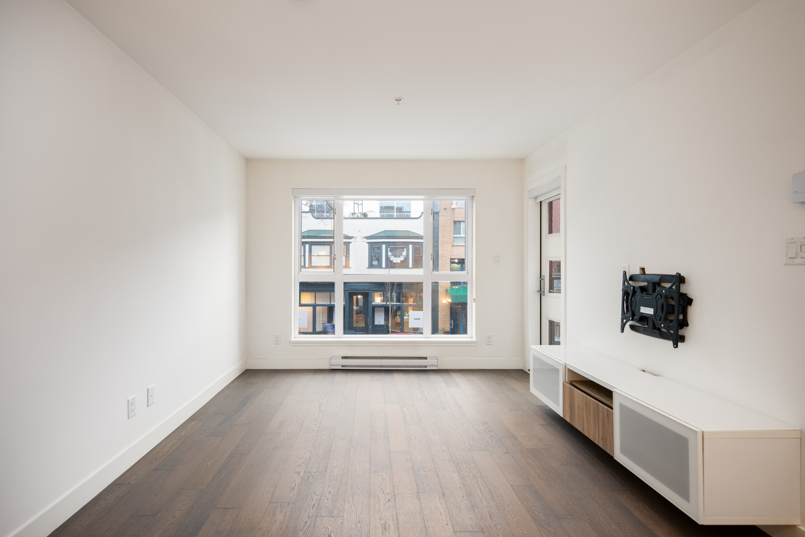Unfurnished living room with white walls, a large window overlooking neighboring buildings, dark wood flooring, and a wall-mounted TV bracket with a low cabinet.