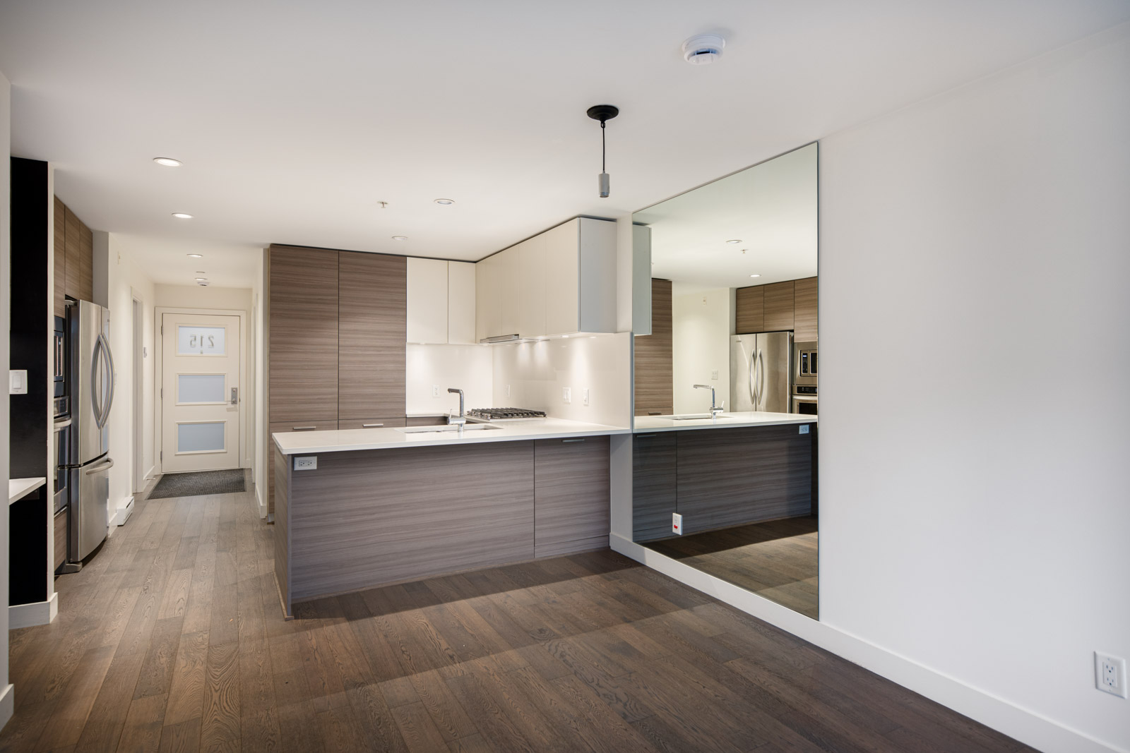 Modern kitchen with wood and white cabinetry, built-in appliances, a large wall mirror, and dark hardwood flooring in a bright, open-concept apartment.