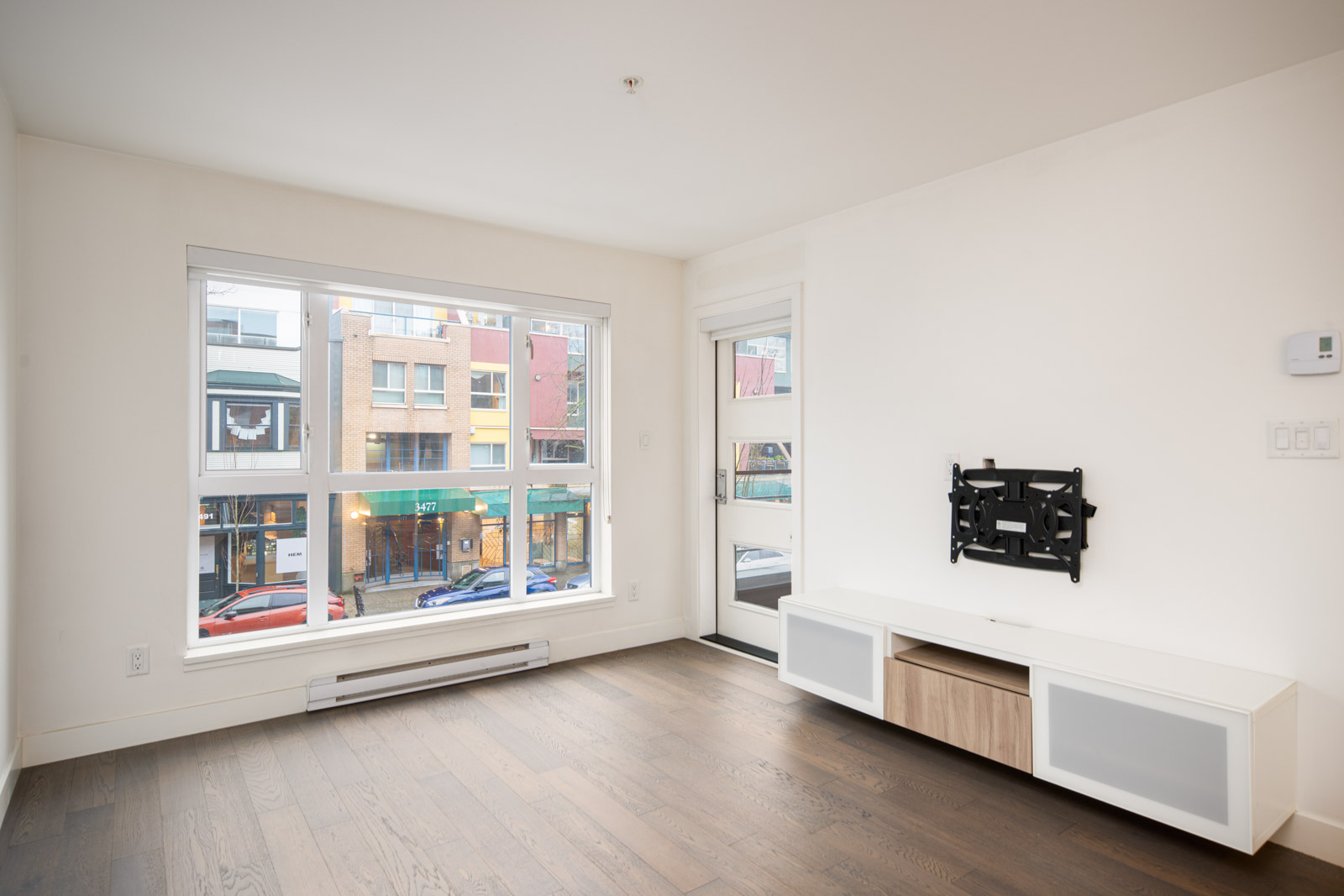 Bright, unfurnished living room with large window, door to balcony, wall-mounted TV bracket, and white media console on hardwood floor. Urban buildings visible outside.