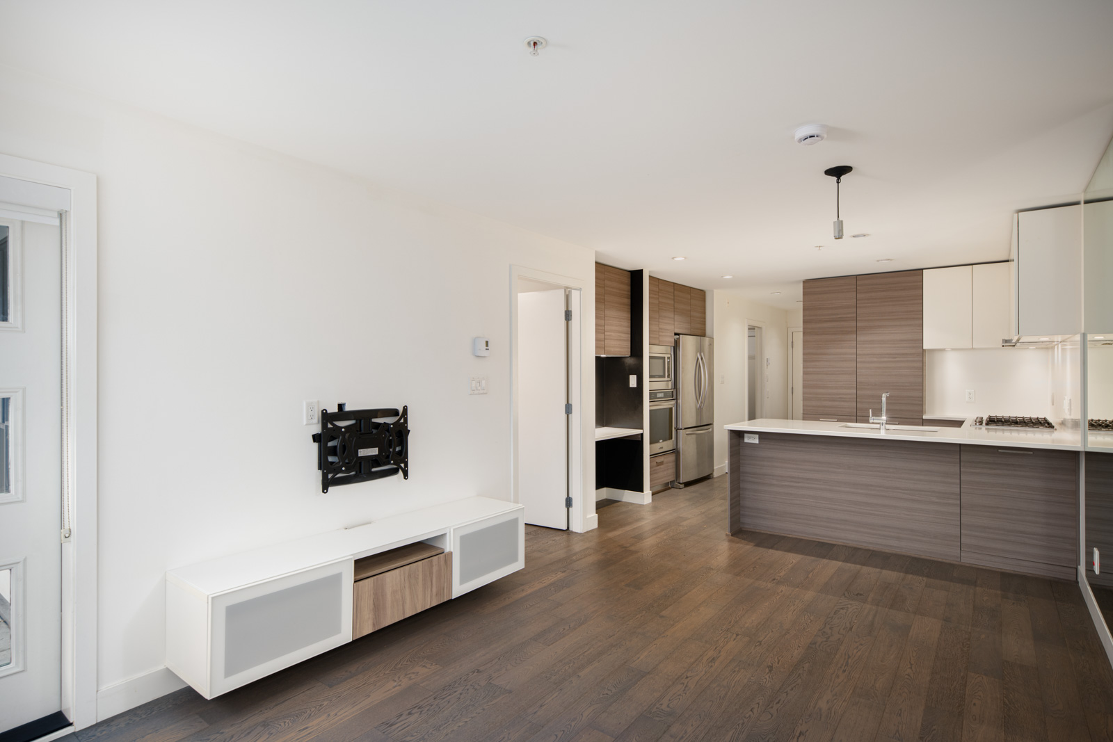 Modern, unfurnished kitchen and living area with wood floors, white walls, built-in cabinets, and stainless steel appliances. TV mount and media console on left wall.