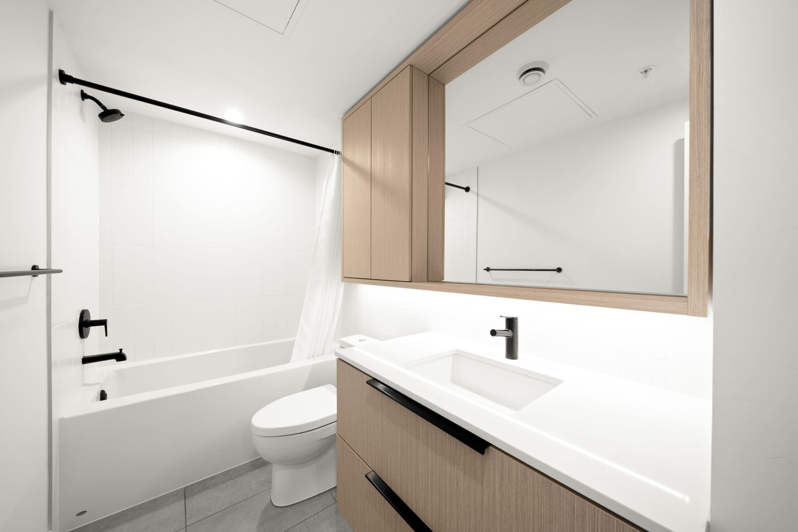 Modern bathroom with white walls, a bathtub with a shower curtain, toilet, wood cabinets, a wide mirror, a sink with a black faucet, and gray tile floor.
